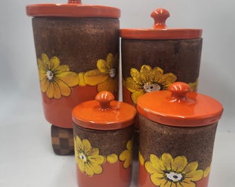 Puede incluir: Un juego de cuatro botes de cerámica con tapas. Los botes son marrones con bases y tapas naranjas. Cada bote presenta un diseño floral amarillo y blanco. Los botes varían en tamaño.