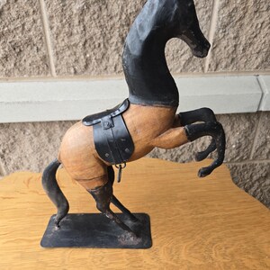 May include: A wooden horse sculpture with a black head and legs, and a saddle. The horse is posed rearing up on its hind legs, mounted on a black rectangular base. The body is a light brown color.