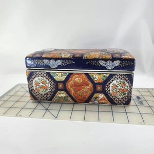 May include: A rectangular ceramic trinket box with a hinged lid. The box features a dark blue base with ornate, colorful patterns of flowers and dragons. The lid is also decorated with similar patterns and floral accents.
