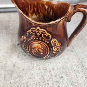 Puede incluir: Un pequeño jarro de cerámica marrón con asa y pico vertedor. El jarro presenta un diseño decorativo en relieve con un medallón central. El esmalte tiene un acabado moteado y brillante, y la estética general es vintage.