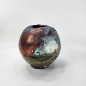 May include: A small, spherical ceramic vase with a dark, iridescent finish. The vase has a narrow opening at the top and a slightly flared base. The colours include shades of brown, blue, and green, creating a unique, textured appearance.