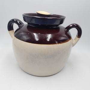 Könnte beinhalten: Ein brauner und weißer Keramiktopf mit Deckel. Der Topf hat zwei Henkel und eine braune Glasur auf der oberen Hälfte. Die untere Hälfte ist weiß.