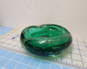 May include: A round, emerald green glass ashtray with a bubbly texture. The ashtray has a unique, organic shape and is made of thick, transparent glass. The ashtray is sitting on a blue and white grid surface.
