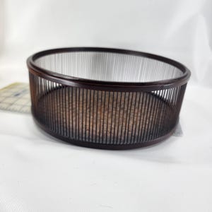 Puede incluir: Una cesta redonda de color marrón oscuro con listones verticales y un borde sólido. La cesta tiene un aspecto natural y tejido y se encuentra sobre una superficie blanca. La cesta probablemente se utiliza para almacenamiento o exhibición.