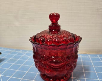 Puede incluir: Un bombonera de cristal rojo rubí con tapa. El plato tiene una base redonda y un patrón decorativo de círculos y espirales. La tapa tiene un pomo redondo en la parte superior. El plato está sobre una superficie azul.