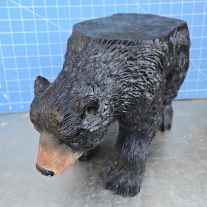 May include: Black bear-shaped stool with a flat top, made of wood and painted black. The bear is in a crouching position with its head down.