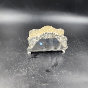 May include: Silver napkin holder with a decorative floral design. The holder has a scalloped edge and three small feet.