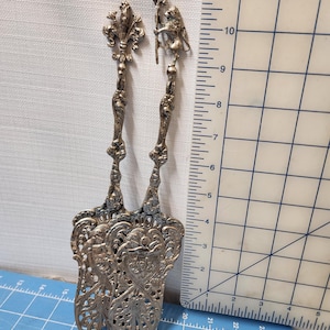 May include: Two ornate silver serving utensils. One features a fleur-de-lis design, the other a lion holding a staff. The utensils are approximately 10 inches long and have intricate, openwork patterns on the serving ends.