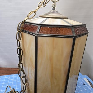 Könnte beinhalten: Eine beige und braune Buntglas-Pendelleuchte mit einer Messingkette.
