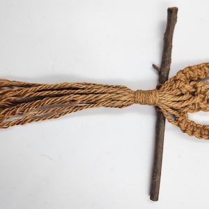 May include: A brown macrame wall hanging with a natural twig attached to the bottom. The macrame is made with a thick, natural fiber rope.