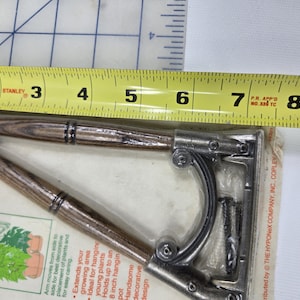 May include: A decorative plant bracket with a wood-grain finish and metal accents. The bracket is approximately 8 inches long, designed for hanging plants. The packaging includes text that reads "For Hanging Plants."