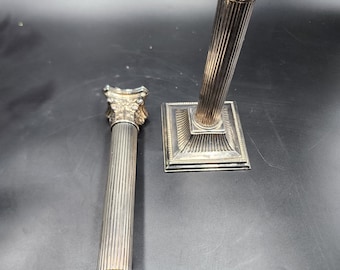 May include: Two silver candlestick holders in the shape of columns with fluted details. The holders are on a black surface.