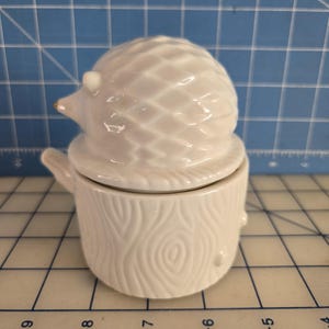 May include: White ceramic hedgehog figurine with a detailed textured design. The hedgehog is sitting on a white ceramic tree stump with a wood grain pattern.