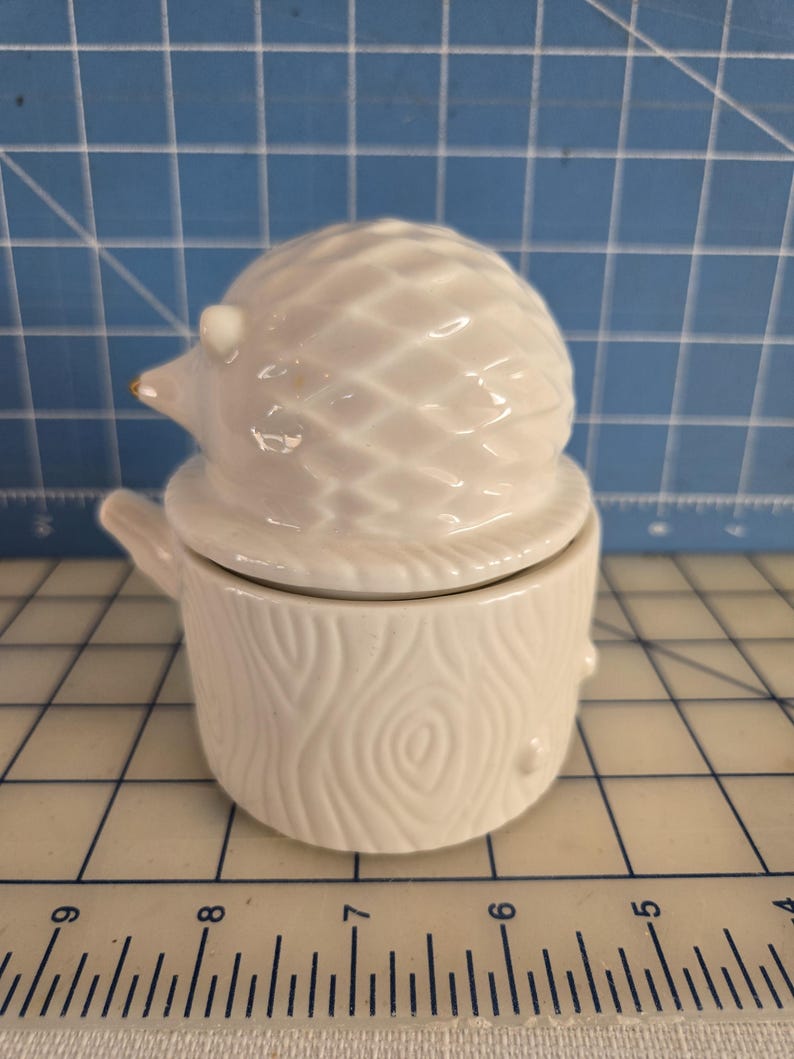 May include: White ceramic hedgehog figurine with a detailed textured design. The hedgehog is sitting on a white ceramic tree stump with a wood grain pattern.