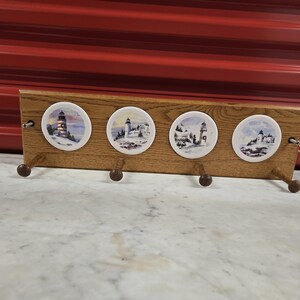 May include: Wooden wall-mounted coat rack with four ceramic lighthouse-themed circular designs. Each design features a different lighthouse scene. The rack has four wooden pegs for hanging items.