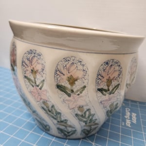 Könnte beinhalten: Cremefarbener Keramik-Blumentopf mit breitem, geschwungenem Rand. Der Topf ist mit sich wiederholenden Blumenmustern in Rosa, Grün und Blau verziert. Die stilisierten Blumen sind vor einem cremefarbenen Hintergrund angeordnet.