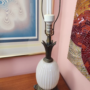 Puede incluir: Una lámpara de mesa de cerámica blanca con una base con forma de piña y una pantalla de metal dorado. La lámpara tiene un diseño acanalado blanco y un remate de metal dorado. La lámpara está iluminada con una bombilla fluorescente compacta.