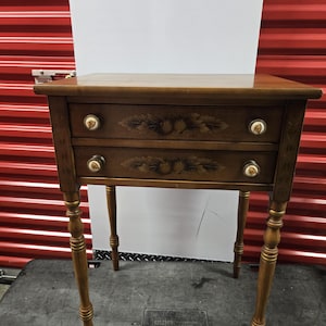 May include: A small, antique wooden side table with two drawers. The table is brown with decorative floral carvings on the drawer fronts and gold accents on the legs. The drawer pulls are white with gold trim. The table is approximately 24 inches tall.