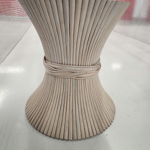 May include: A light beige, hourglass-shaped side table. The table is made from vertical, thin wooden slats, bound with a woven band around the middle. The table has a flared top and base, creating a unique, decorative design.