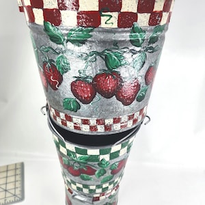 May include: Two metal strawberry-themed buckets stacked together. The buckets are silver with hand-painted red strawberries and green leaves. The rims feature a red and white checkered pattern. One bucket has the number 2.