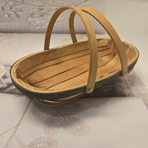 May include: An oval-shaped wooden basket featuring a light natural wood interior and a dark green painted exterior rim. The basket includes two arched wooden handles that are secured to the sides with small metal rivets, creating a sturdy structure.