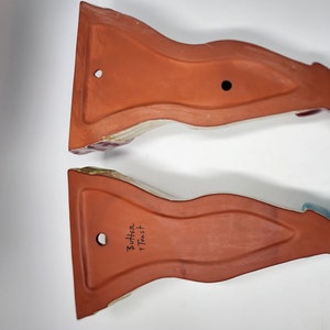 May include: Two terracotta wall-mounted fish-shaped dishes with a glazed finish. The bottom of each dish has a hole for hanging and the text "Butter + Toast" is written on one of the dishes.