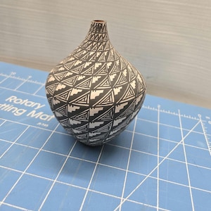 May include: A black and white ceramic vase with a geometric pattern. The vase has a rounded body and a narrow neck. The design features repeating triangles and stepped shapes. The vase is displayed on a blue cutting mat.