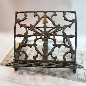 May include: Ornate bronze-colored metal book stand with an openwork design. The stand features a rectangular back with a decorative pattern and a small shelf at the bottom. The stand is supported by two legs and is designed to hold books or other items.