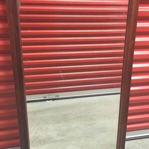 May include: A rectangular mirror with a dark wood frame. The mirror is reflecting a red metal garage door.