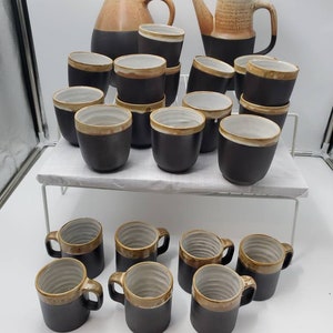May include: A set of 15 ceramic mugs and two ceramic teapots in a brown and tan glaze. The mugs have a brown exterior and a tan interior. The teapots have a tan exterior and a brown interior.