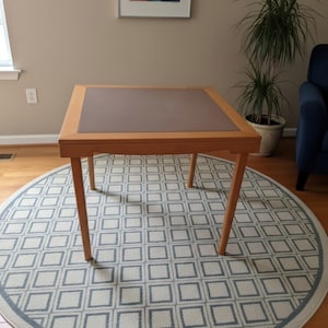 May include: A square wooden table featuring a smooth, light-toned frame and a flat, matte-finished center panel in a muted taupe color. The table is supported by four straight, tapered legs that extend outward from the corners to the floor.