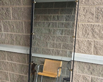 May include: A tall, rectangular mirror with a black frame and gold-colored accents. The mirror is leaning against a brick wall. The frame has a simple, modern design. The mirror reflects the surrounding environment.
