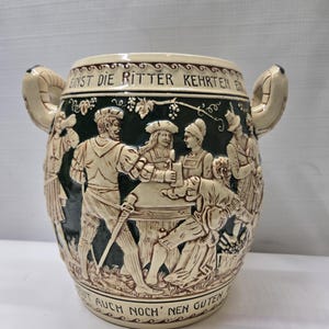 Op de afbeelding: Een steengoed kruik met een groen en bruin ontwerp met een scène van mensen die rond een tafel zijn verzameld. De kruik heeft twee handvatten en de tekst "Ein St die Ritter Kehrten E... Auch Noch' nen Guten" staat rond de boven- en onderkant.