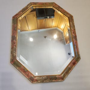 Puede incluir: Espejo octogonal con marco estampado floral en tonos rosa, verde y dorado. El espejo refleja una habitación con gabinetes de madera y luces brillantes, creando un acento decorativo de pared.