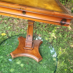 May include: A wooden table with a rectangular top and a pedestal base. The tabletop is a warm brown with a lighter center, and the base is a darker brown. The table is supported by a clear, round glass top.