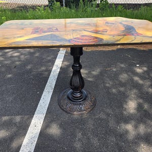 Op de afbeelding: Een rechthoekige houten tafel met een beschilderd blad met een kleurrijke scène. De tafel heeft een zwarte, sierlijke voet. De afmetingen van de tafel zijn ongeveer 122 cm lang, 76 cm breed en 76 cm hoog.