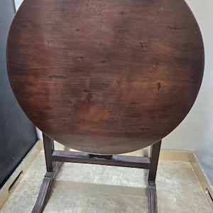Puede incluir: Una mesa redonda de madera marrón oscuro con base plegable y patas talladas. La mesa está sobre una superficie gris.