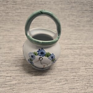 May include: A small white ceramic basket with a rounded body and a single arched handle painted in a muted green. The front features a hand-painted design of a stylized bird surrounded by a wreath of blue flowers and green leaves.