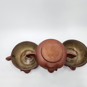 May include: A set of three brown ceramic teaware pieces. The set includes a teapot and two matching cups. The teapot is centered, with the cups on either side. The teaware has a rustic, earthy aesthetic.