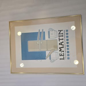May include: A framed poster with a blue background and a white illustration of a coffee pot, cups, and croissants. The text "LE MATIN" is in large, bold letters and the text "9+ CAFE & CROISSANT CAFE CHEZ PANISSE" is in smaller letters below.