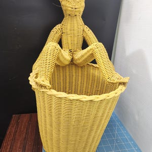 May include: A yellow wicker basket with a woven figure of an alien sitting on the rim. The alien has large eyes and is made of the same woven material as the basket. The basket is cylindrical and has a wide opening.
