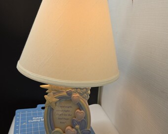 May include: A decorative bedside lamp with a cream-colored lampshade and a base featuring a pastel rainbow, stars, and hearts. The base has the text "Good night, sleep tight, Don't let the bed bugs bite."