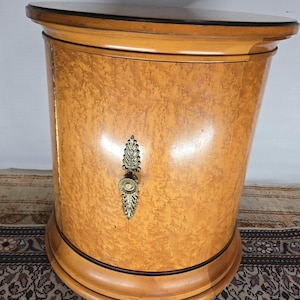May include: A round wooden side table with a single door and a brass handle. The table has a light wood finish with a darker wood trim around the top and bottom.