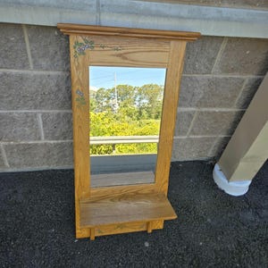 Puede incluir: Un espejo rectangular de madera con una repisa. El espejo está enmarcado en madera marrón claro y presenta una pequeña repisa en la parte inferior. La parte superior del marco tiene un borde decorativo y la esquina superior izquierda tiene un diseño floral pintado.