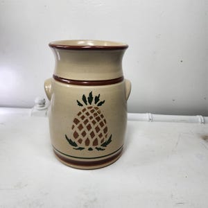 May include: Tan ceramic jar with a brown pineapple design. The jar has a wide opening, two small handles, and brown trim around the top and bottom. The pineapple is brown with green leaves.