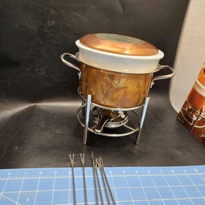 May include: A copper fondue pot with a white ceramic insert and a copper lid. The pot sits on a chrome stand with a burner. Several fondue forks are in front of the pot, ready for dipping.