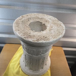May include: A cylindrical stone pedestal with a rough, textured top surface featuring a central circular opening. The body of the pedestal is decorated with vertical fluted columns and carved leaf motifs around the upper rim in a light gray color.