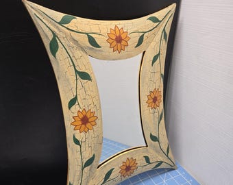 Puede incluir: Un espejo decorativo con un marco crema con acabado craquelado. El marco tiene una forma curva única y está adornado con girasoles amarillos pintados y hojas verdes. El espejo es un artículo de decoración del hogar.