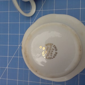 May include: A white teacup saucer with a gold rim and the text "WEST END POTTERY LIVERPOOL OHIO" in gold lettering. The saucer is on a blue grid surface. A portion of a white teacup is visible.