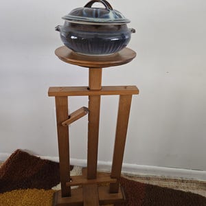 Op de afbeelding: Een houten standaard met een ronde bovenkant, waarop een blauwe en bruine keramische pot met deksel staat. De standaard heeft een eenvoudig ontwerp met vier poten en een kruisvormige basis.